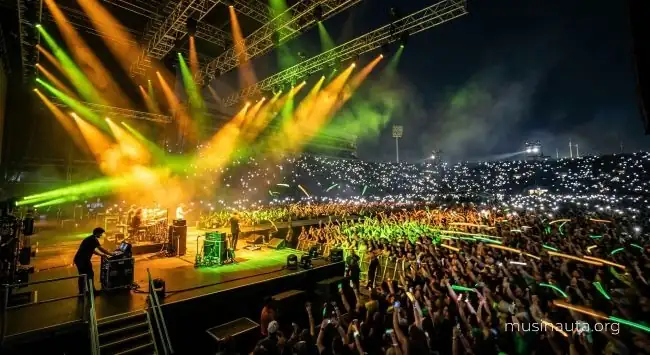 Concierto nocturno al aire libre con luces de escenario y multitud iluminada por pantallas de teléfono