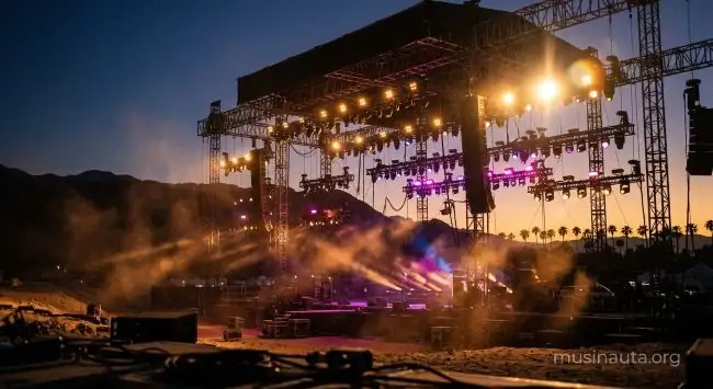 Escenario de festival de música en el desierto con estructuras de luces encendidas al atardecer y polvo en el aire