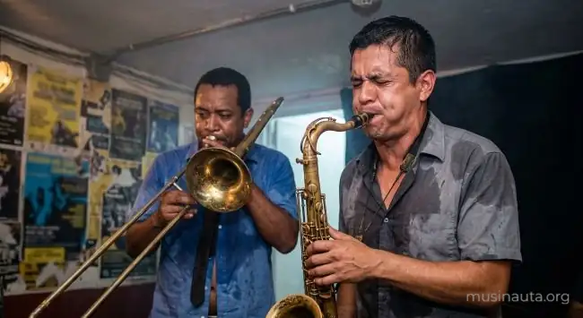 Saxofonista y trombonista tocando con los ojos cerrados y las camisas empapadas en el escenario bajo de un club pequeño