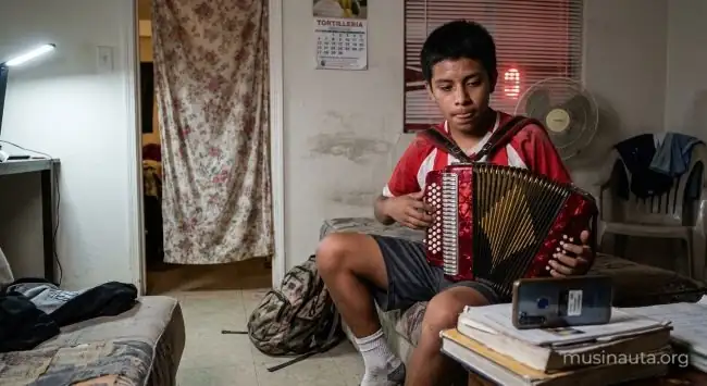 Adolescente aprende a tocar un acordeón de botones siguiendo un tutorial en el celular dentro de un departamento